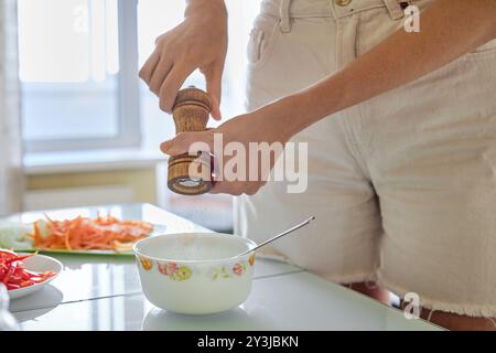 Una persona sta macinando il pepe fresco in un recipiente stando in piedi davanti a un luminoso bancone della cucina. Le verdure tritate sono disposte sul tavolo, mostrando una V Foto Stock