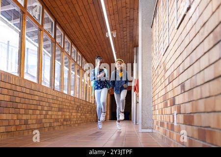 Correndo nel corridoio della scuola, due ragazze afroamericane con zaini che sorridono e si divertono Foto Stock