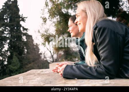 Due ragazze si appoggiano su una vecchia ringhiera in pietra e si godono la vista. Donne affascinanti in abiti informali autunnali chiacchierano nel parco sulla piattaforma di osservazione. Foto Stock