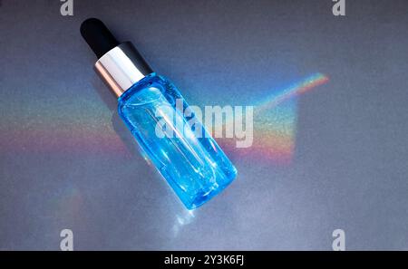 Un primo piano di una beuta per siero blu riflette un arcobaleno su uno sfondo scuro. Un modello di un prodotto cosmetico. Un flacone di vetro con pipetta e idratazione Foto Stock