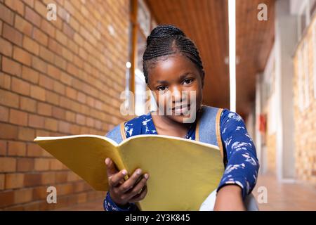 Con un taccuino, una ragazza afroamericana che sorride e legge nel corridoio della scuola, indossa uno zaino Foto Stock