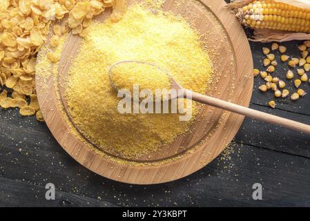 Immagine rustica con un cucchiaio di legno pieno di farina di mais in cima a un pannello di legno, circondato da fiocchi di mais, grani e pannocchie di mais, su un tavolo di legno nero Foto Stock