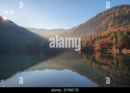 Mattina di ottobre con il sole che splende sulle colline con la colorata foresta autunnale e la sua riflessione sul lago Alpsee, a Fussen, Baviera, Germania, Europa Foto Stock