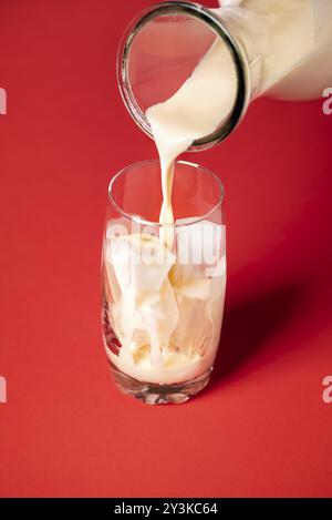 Fare horchata mocktail contesto. Versare il latte di horchata in un bicchiere con cubetti di ghiaccio su fondo rosso. Serve drink estivi messicani. Bevanda fatta in casa Foto Stock
