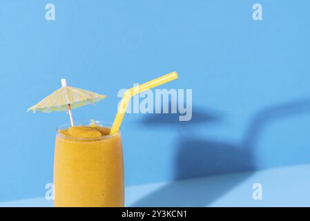 Frullato di mango in un bicchiere con un ombrello di paglia e bevanda su sfondo blu. Primo piano del cocktail di mango. Bevanda tropicale. Bevanda gialla estiva Foto Stock