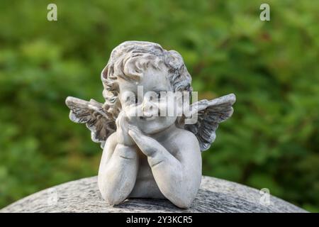 Una piccola statua d'angelo come decorazione funeraria in un cimitero. L'immagine ha uno spazio vuoto per il testo. La statua ha uno sfondo verde estivo. Foto Stock