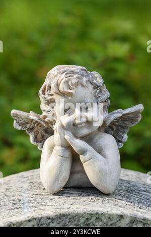 Una piccola statua d'angelo come decorazione funeraria in un cimitero. L'immagine ha uno spazio vuoto per il testo. La statua ha uno sfondo verde estivo. Foto Stock