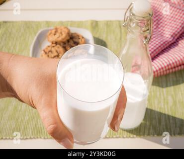 Fotografia di cibo con un bicchiere di latte tenuto in mano a una donna, il biberon di latte e i biscotti stesi sul tavolo sullo sfondo Foto Stock