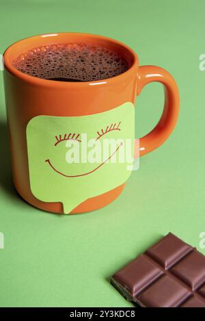 Il caffè caldo in una tazza di colore arancione e una divertente Smiley face nota adesiva su un tavolo verde accanto a un grande pezzo di cioccolato Foto Stock