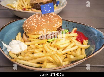 Fotografia gastronomica con un delizioso hamburger grande, patatine fritte, maionese, ketchup e una piccola bandiera dell'Unione europea in cima Foto Stock