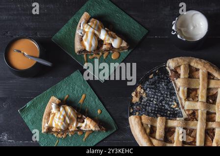 Vista dall'alto di un tavolo con una torta di mele intera e due fette su tovaglioli, decorato con caramello e panna montata. Dolci fatti in casa Foto Stock