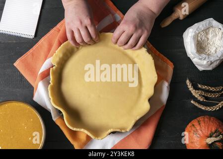 Donna che lavora con le mani un impasto per torta in un vassoio, su un tavolo da cucina, circondato da ingredienti per la torta di zucca e un quaderno. Torte tradizionali da forno Foto Stock