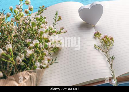 Piccoli fiori bianchi e un cuore di legno su pagine vuote di quaderni, su sfondo blu. Primo piano. Cornice di San Valentino. Contesto per la pianificazione di matrimoni Foto Stock