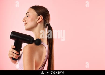 Donna che utilizza un massaggiatore a percussione per alleviare i muscoli trapezoidali su sfondo rosa. Spazio per il testo Foto Stock