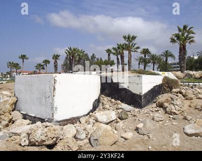 Bunker abbandonati in cemento dipinto di bianco a paphos cipro, risalenti all'epoca della guerra civile, con rocce sulla spiaggia accanto a palme e hotel Foto Stock