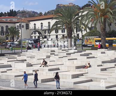 Funchal, Madiera, Portogallo, 15 marzo 2019: Turisti seduti e passanti sui gradini concreti del porto turistico di funchal di fronte alla città vecchia con il suo Foto Stock