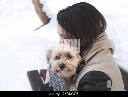 Giovane donna bruna seduta su una panchina, circondata dalla neve, che tiene con affetto il suo cane Havanese Bichon, per proteggerlo dal freddo Foto Stock
