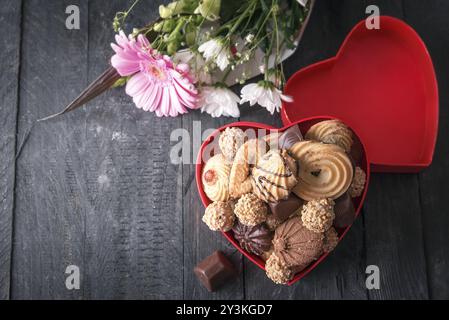 Vista ad alto angolo con una scatola rossa a forma di cuore piena di biscotti e cioccolatini e un bouquet di crisantemo sullo sfondo, su un tavolo di legno nero Foto Stock