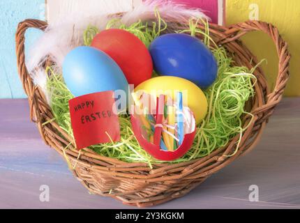 Cestino di vimini pieno di uova dipinte e guscio d'uovo con note di carta arrotolate e uno con le parole "buona pasqua", su un colorato sfondo di legno Foto Stock