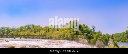 Ampia vista con il grande flusso del fiume Reno e la sua cascata, un ponte che lo attraversa e un piccolo palazzo che lo affaccia dalla cima di una collina Foto Stock