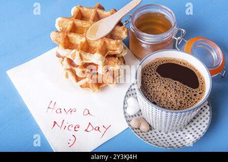 Tazza di caffè con zucchero a forma di cuore, waffle fatti in casa con miele e un tovagliolo con un bel messaggio di giornata, su un tavolo di legno blu Foto Stock