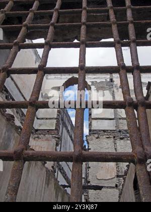 Vista attraverso barre di ferro arrugginite su una finestra di un edificio crollato con pareti sbriciolate e aperto verso il cielo Foto Stock