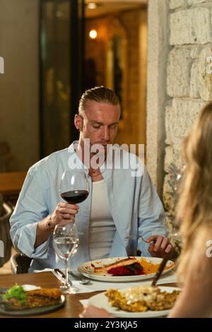 Un uomo assaggia attentamente il suo bicchiere di vino rosso mentre cena in un elegante ristorante all'aperto, assaporando i sapori Foto Stock