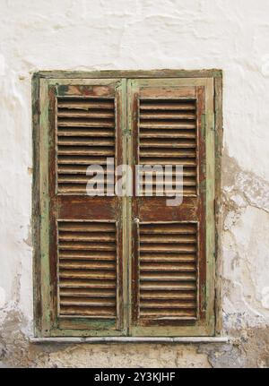 Antiche persiane chiuse dipinte di verde marrone in una parete bianca con cemento incrinato e intonaco Foto Stock