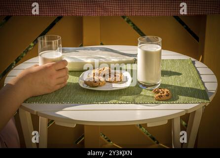 Un bicchiere di latte tenuto in mano a una donna e un piatto con biscotti, su un tavolo rotondo bianco, in una calda mattinata di sole Foto Stock