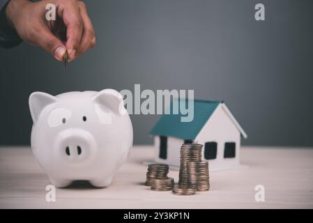 Mettere a mano la moneta nel salvadanaio bianco accanto al modello della casa e a pile di monete. Simboleggia risparmi, crescita finanziaria e investimenti immobiliari. BUDG Foto Stock