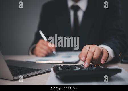 Primo piano di un uomo d'affari con una tuta che lavora alla scrivania. Calcolatrice a pressione manuale, analisi dei documenti. Professionale, focalizzato sui calcoli finanziari. Foto Stock