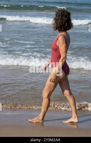 Una donna anziana cammina lungo la riva di una spiaggia sabbiosa, indossando un costume da bagno rosso monopezzo. Foto Stock