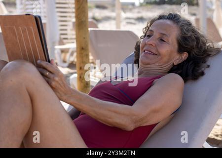 Una donna anziana si sta godendo la sua vacanza al mare, sdraiata su un lettino da spiaggia e leggendo un e-book. Foto Stock