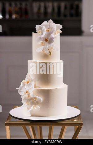 Elegante torta nuziale a tre piani decorata con lussuose orchidee bianche e iniziali oro su un moderno espositore. Foto Stock