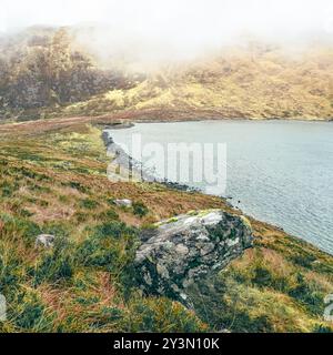 Un tranquillo lago è annidato tra aspre colline, con la nebbia che si libra dolcemente sul paesaggio durante la mattina presto. La scena trasuda calma e naturale Foto Stock