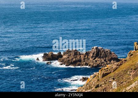 Una vista panoramica della costa rocciosa con onde che si infrangono contro le rocce, circondate da profonde acque blu dell'oceano e un cielo limpido. Foto Stock