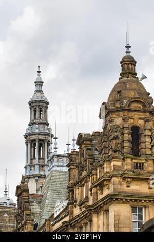 Vista ravvicinata di dettagli architettonici ornati su edifici storici, con un'alta guglia e una cupola con intricate incisioni contro un cielo nuvoloso. Foto Stock