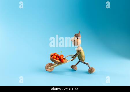 Affascinante statuetta autunnale realizzata con ghiande, con corpo e testa di ghiande, arti ramoschianti, che spingono una carriola ramoschiata piena di bacche rosse, blu Foto Stock