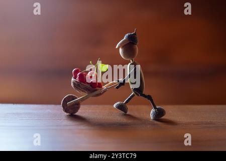 Affascinante statuetta autunnale realizzata con ghiande, con corpo e testa di ghiande, arti ramoschianti, che spingono una carriola ramoschiata piena di bacche rosse, blu Foto Stock