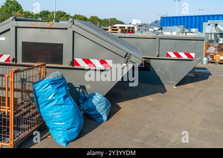 Due grandi contenitori per rifiuti si trovano in un'area industriale sotto cieli limpidi. Nelle vicinanze, accanto ai contenitori sono collocati sacchetti blu dei rifiuti, in attesa di essere prelevati Foto Stock