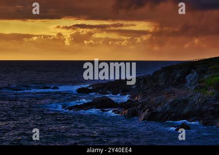 Una spettacolare scena costiera al tramonto, caratterizzata da scogliere rocciose e onde turbolente. Il cielo è pieno di vivaci tonalità arancio e viola, che riflettono il Foto Stock