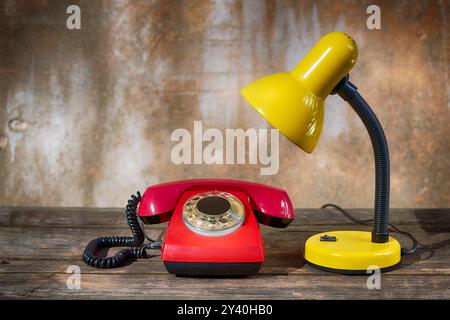 Una lampada da tavolo e un telefono rosso vintage su un vecchio tavolo di legno su uno sfondo squallido Foto Stock