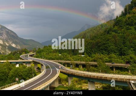 Sotto un ampio skyline, una strada tortuosa attraversa una vegetazione lussureggiante che conduce a maestose montagne. Un brillante arco arcobaleno si innalza aggraziatamente sopra, Foto Stock