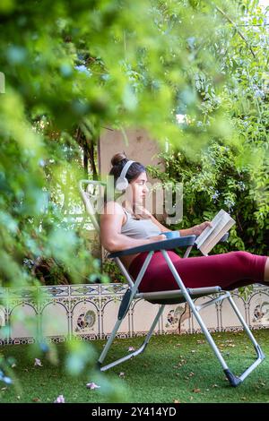 Ragazza seduta su una sedia a sdraio nel suo giardino a leggere un libro e a bere caffè, nelle piante in primo piano fuori fuoco. Foto Stock