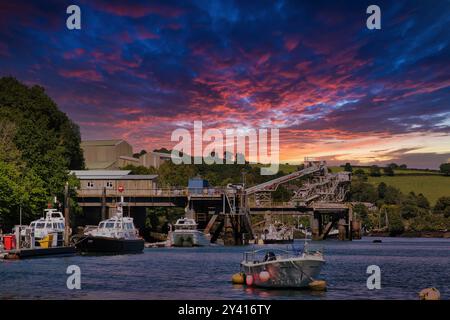 Una vista panoramica di un porto al tramonto, con barche in primo piano e un molo con strutture industriali sullo sfondo. Il cielo è pieno di Foto Stock