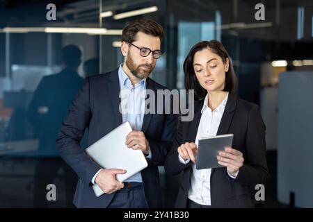 Professionisti aziendali impegnati in discussioni utilizzando tablet in ufficio. Uomo che tiene un notebook mentre una donna dimostra con un tablet, mostrando il lavoro di squadra e la comunicazione in ambiente aziendale Foto Stock