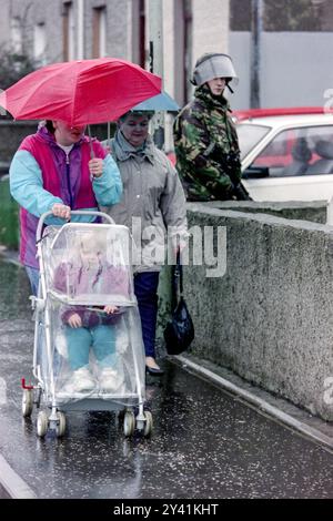 Una famiglia spinge una carrozza in un giorno di pioggia davanti a un soldato britannico che pattuglia l'area di Bogside, il 25 gennaio 1992 a Londonderry, Irlanda del Nord. Le forze di sicurezza hanno intensificato le pattuglie e le perquisizioni nella zona prima del ventesimo anniversario del massacro di Bloody Sunday. Foto Stock