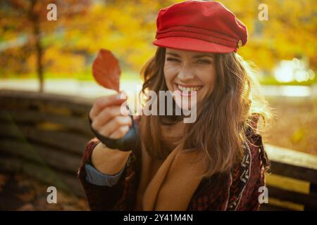 Ciao autunno. Ritratto di donna sorridente alla moda con cappello rosso con foglie autunnali e sciarpa seduta sulla panchina nel parco cittadino. Foto Stock