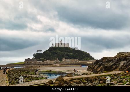 Una vista panoramica di un paesaggio costiero caratterizzato da un castello su una collina, circondato da coste rocciose e una spiaggia di sabbia. La gente cammina lungo il sentiero e. Foto Stock