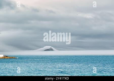 Un tranquillo paesaggio marino caratterizzato da calme acque turchesi sotto un cielo nuvoloso. In lontananza, una piccola isola è parzialmente oscurata dalla nebbia, creando un mistico Foto Stock